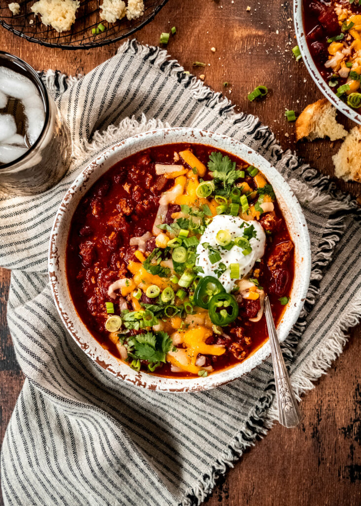 bowl of turkey chili garnished with shredded cheese, cilantro, sliced green onion and sour cream.  The bowl is sitting on a striped napkin on a wood table next to a water glass. 