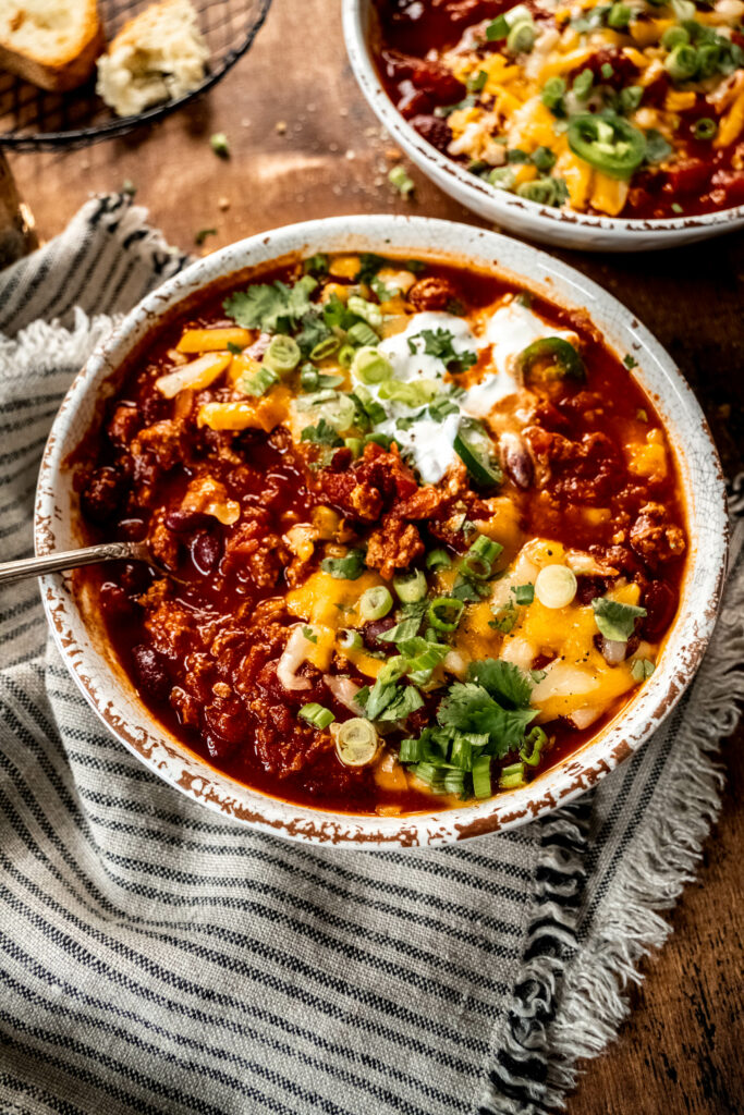 bowl of turkey chili garnished with shredded cheese, cilantro, sliced green onion and sour cream.  The bowl is sitting on a striped napkin on a wood table