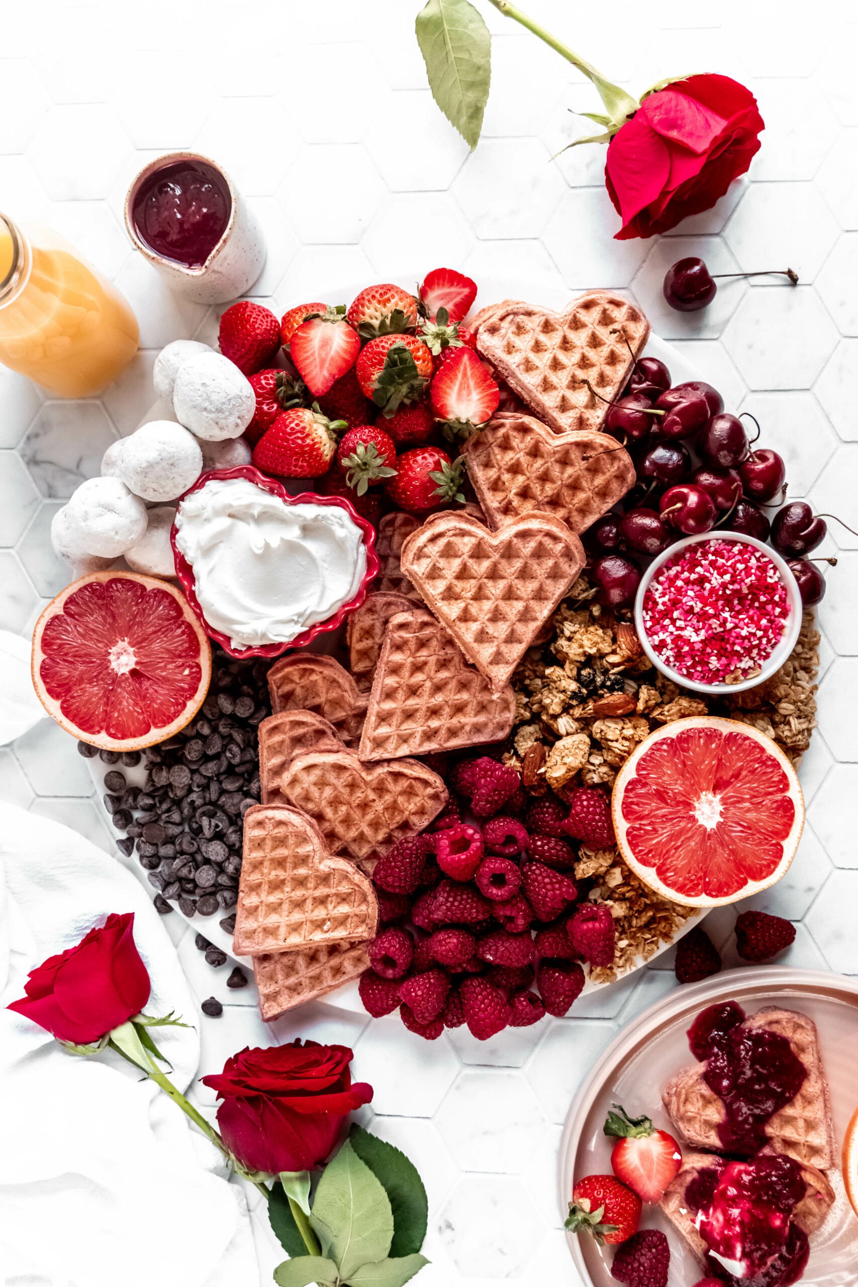 full overhead shot of  valentines day breakfast board with heart shaped waffles, raspberries, strawberries, grapefruit, chocolate chips, granola, and shipped cream on hexagon marble background with red roses on the counter  