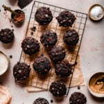 banana chocolate chip muffins on a wire cooling rack surrounded by chocolate chips, a bowl of cocoa powder and neutral linens