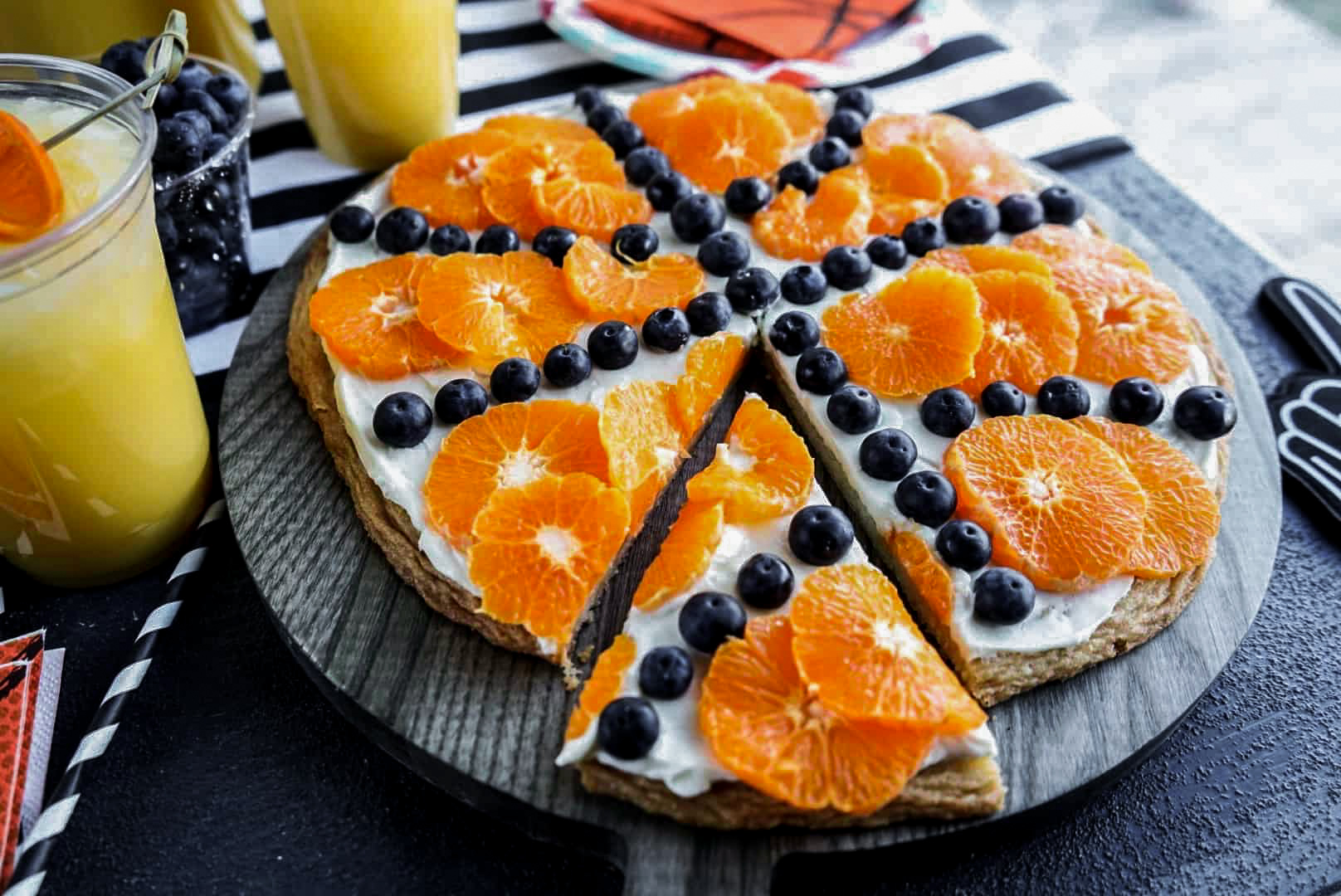 Sliced Sugar Cookie Fruit Pizza decorated like a basketball with cream cheese frosting, mandarin orange slices, and blueberries, served on a round wooden board with basketball-themed party decor.
