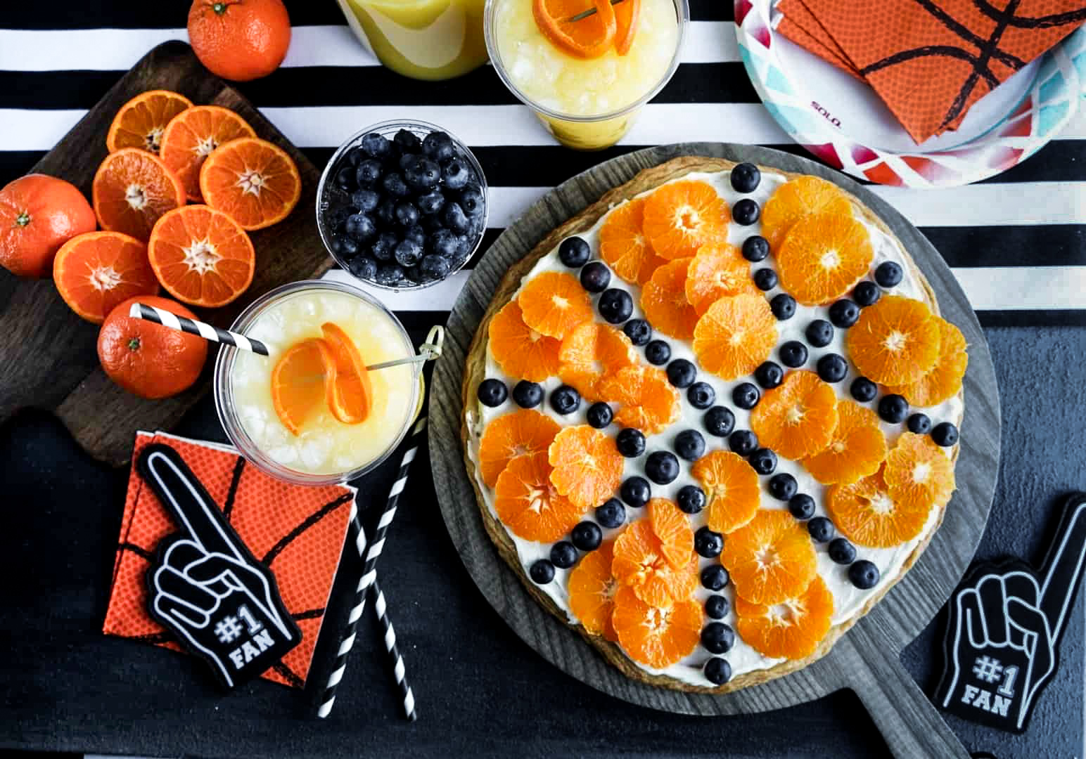 basketball fruit pizza Overhead view of a Sugar Cookie Fruit Pizza decorated like a basketball with cream cheese frosting, mandarin orange slices, and blueberries arranged on a round cookie crust, surrounded by basketball-themed party decor.