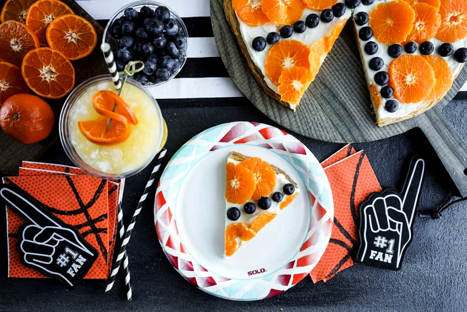 basketball fruit pizza Overhead view of a slice of Sugar Cookie Fruit Pizza on a plate, topped with cream cheese frosting, mandarin oranges, and blueberries, styled with basketball-themed napkins, drinks, and party decor.