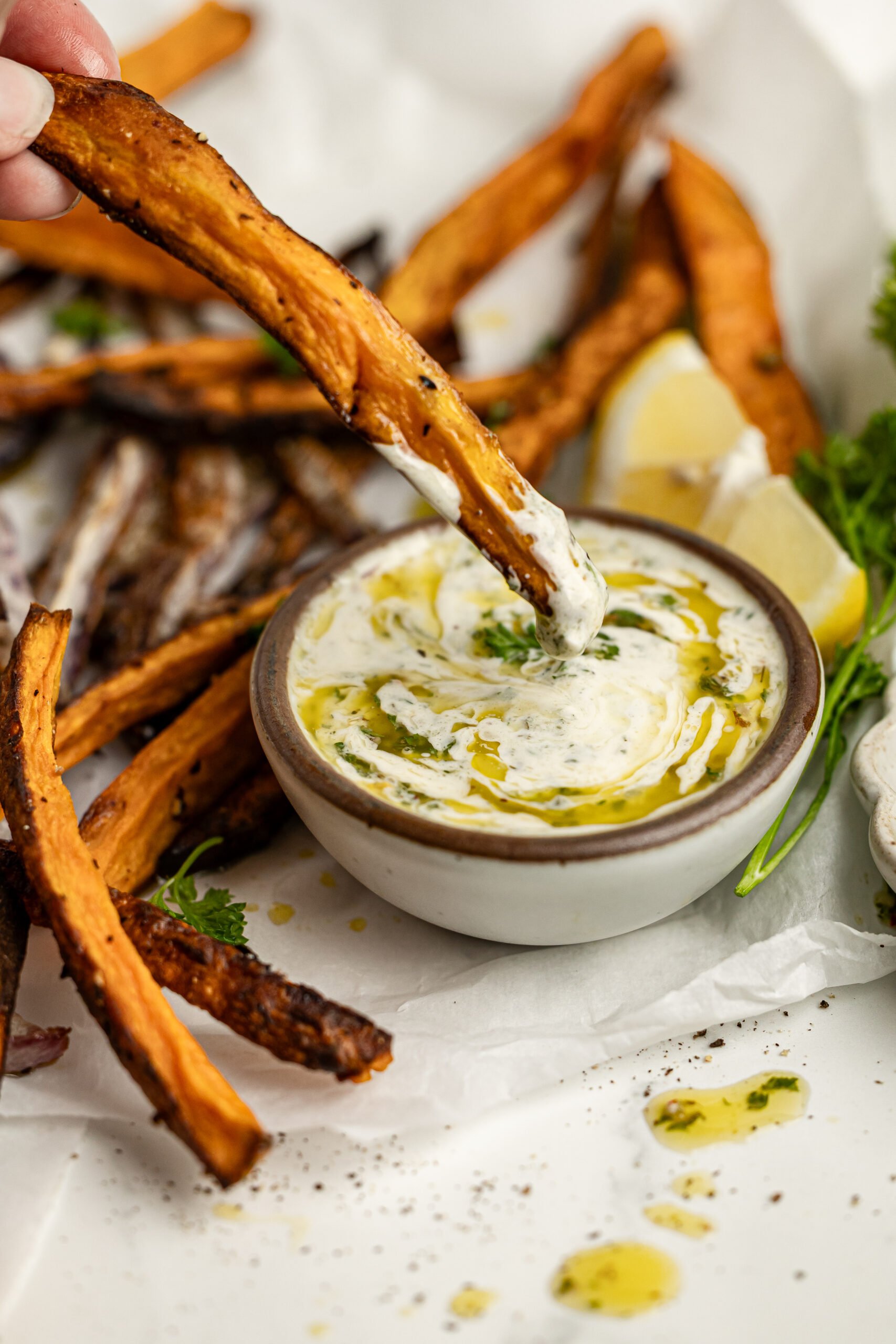 a side shot of a french fry being dipped in chimichurri aioli in a small bowl.  The bowl is  surrounded by crispy french fries, a lemon wedge and fresh parsley