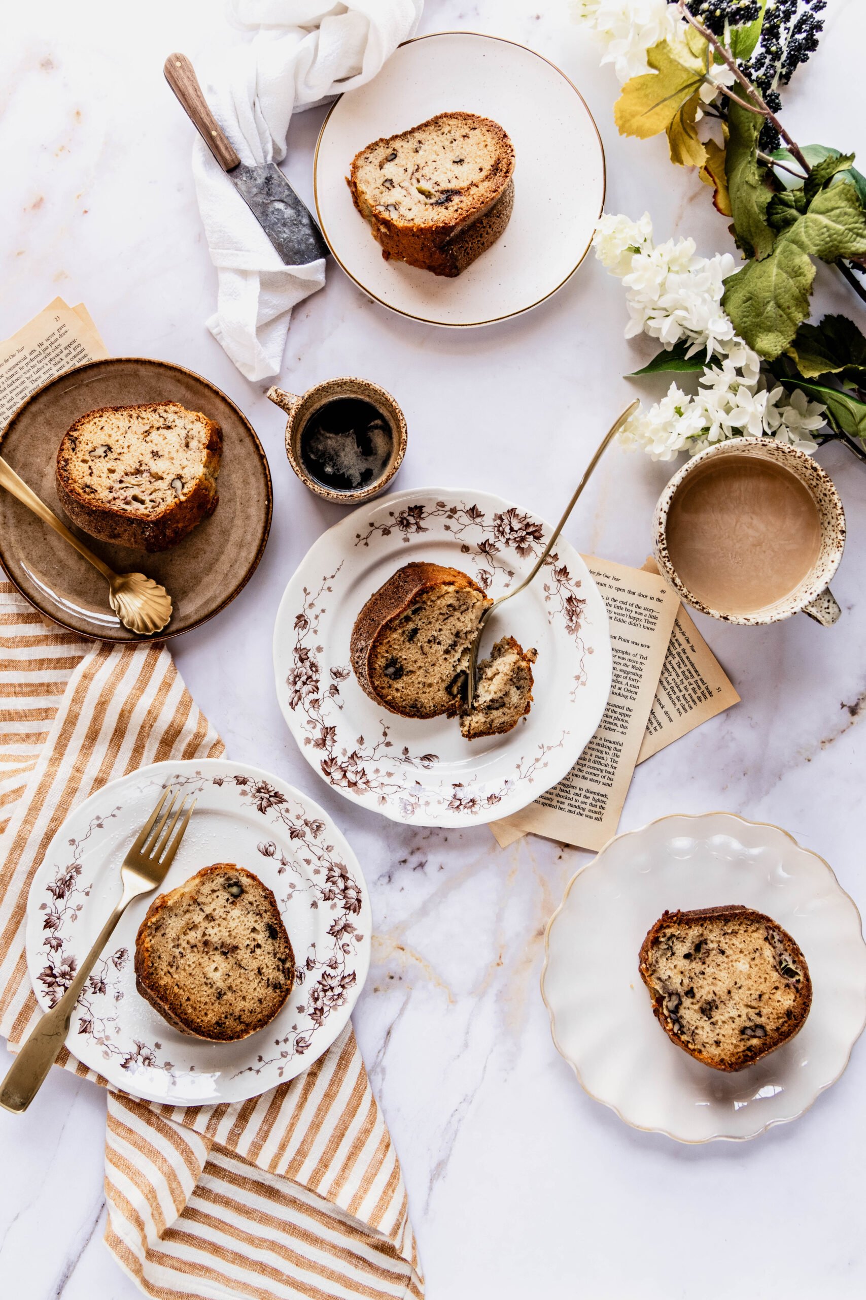 Banana nut bread bundt cake sliced on  five round plates on a white marble countertop. There's two cups of coffee next to the plates and a yellow and white striped towel on the counter. 