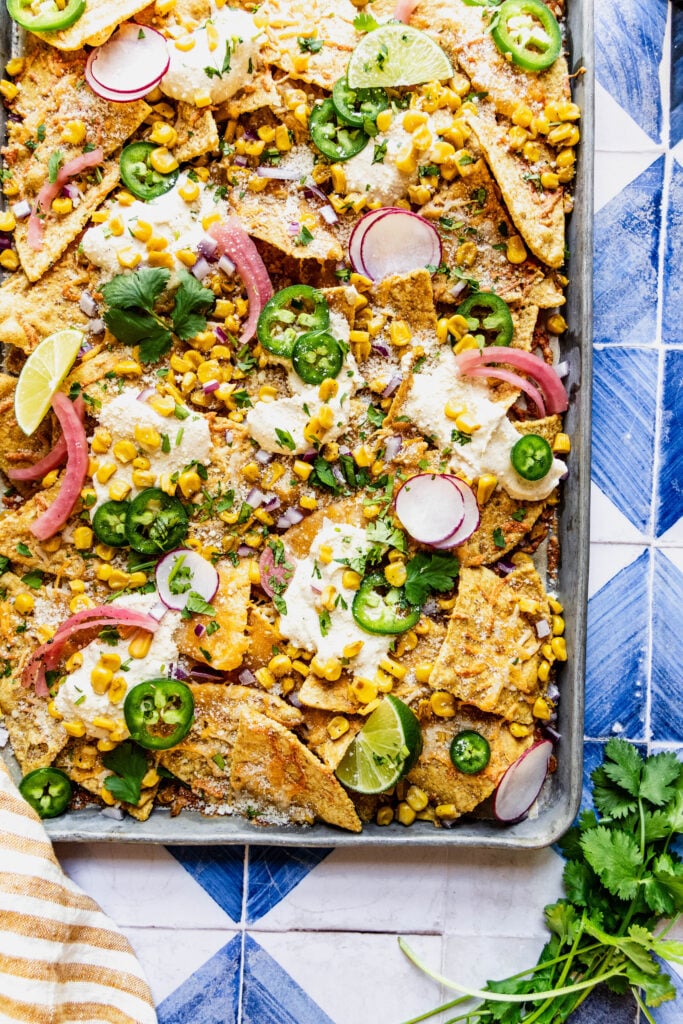 close up overhead shot of elote nachos garnished with jalapeno slices, cilantro, pickled onions and lime wedges.  The nachos are on a sheet pan on a blue and white tile background 