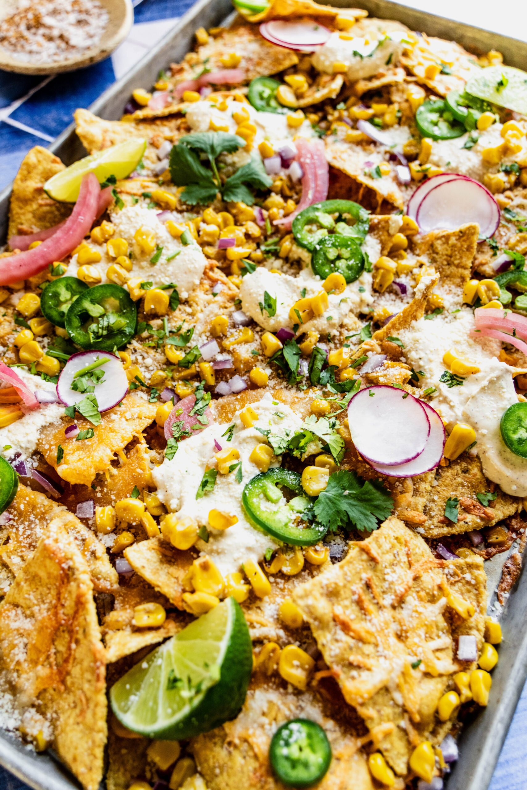 close up side shot of elote nachos garnished with jalapeno slices, cilantro, pickled onions and lime wedges.  The nachos are on a sheet pan on a blue and white tile background 