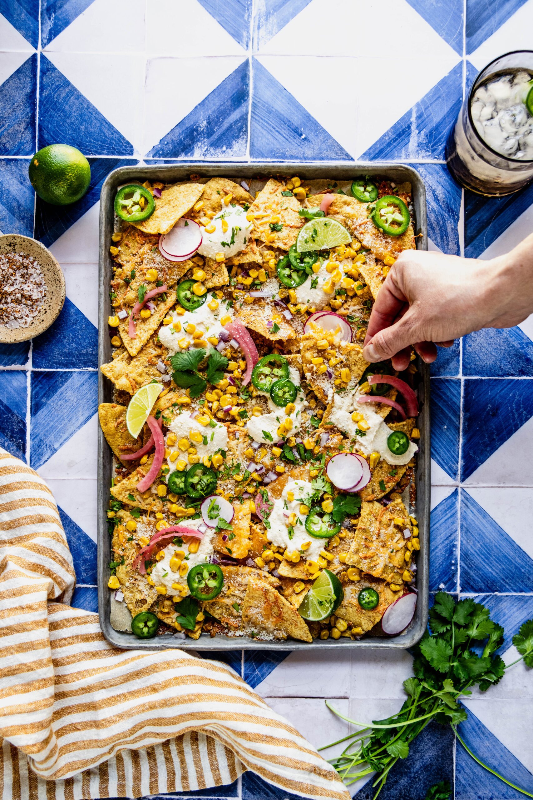 overhead shot of a hand reaching for a chip on the elote nachos garnished with jalapeno slices, cilantro, pickled onions and lime wedges.  The nachos are on a sheet pan on a blue and white tile background 