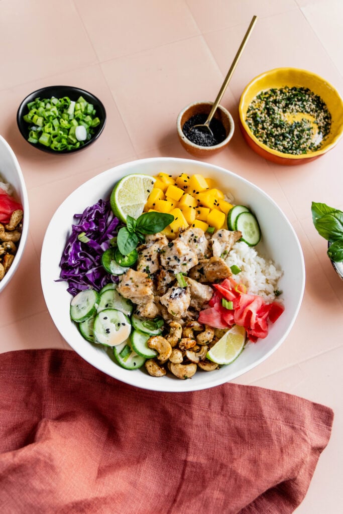 Alt text:
Close-up of a colorful chicken poke bowl featuring grilled miso chicken, sushi rice, diced mango, sliced cucumber, shredded purple cabbage, pickled ginger, furikake cashews, lime wedges, and fresh herbs, with small bowls of scallions, black sesame seeds, and dressing in the background.
