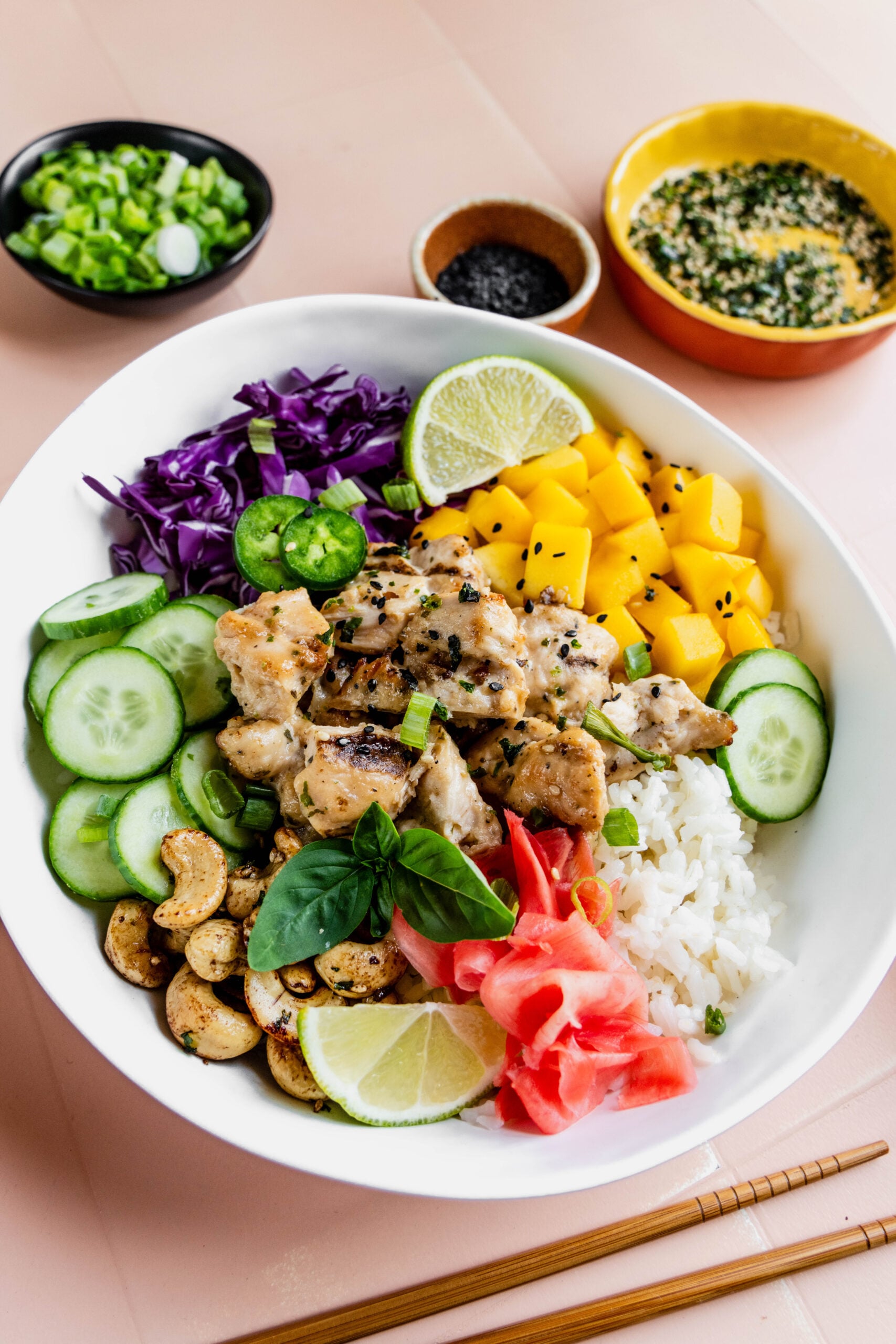 Alt text:
Close-up of a colorful chicken poke bowl featuring grilled miso chicken, sushi rice, diced mango, sliced cucumber, shredded purple cabbage, pickled ginger, furikake cashews, lime wedges, and fresh herbs, with small bowls of scallions, black sesame seeds, and dressing in the background.