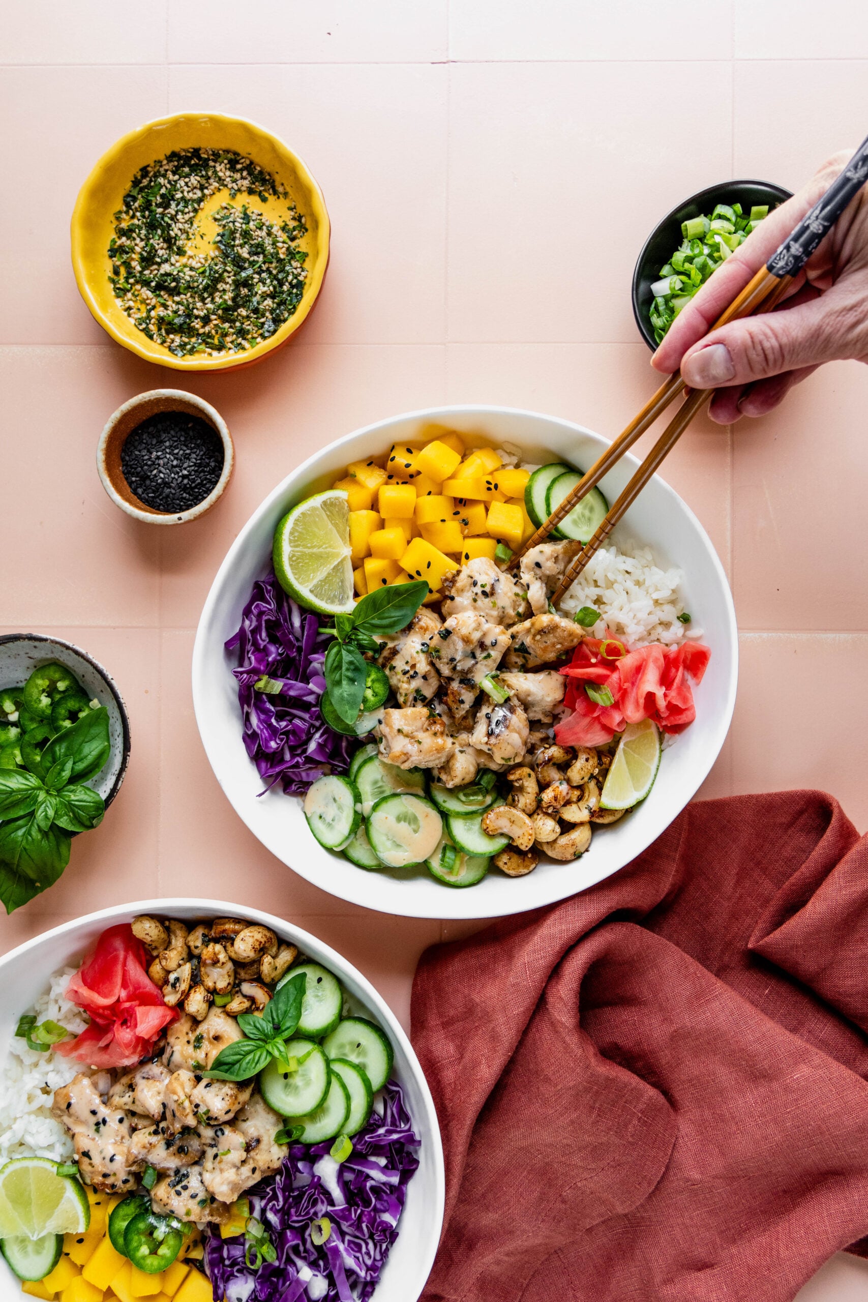 Alt text:
Overhead view of a vibrant chicken poke bowl filled with sushi rice, miso chicken, diced mango, cucumber slices, shredded purple cabbage, furikake cashews, pickled ginger, and lime wedges, surrounded by small bowls of toppings and sauces on a peach-colored surface. A hand with chopsticks reaches into one of the chicken poke bowls.