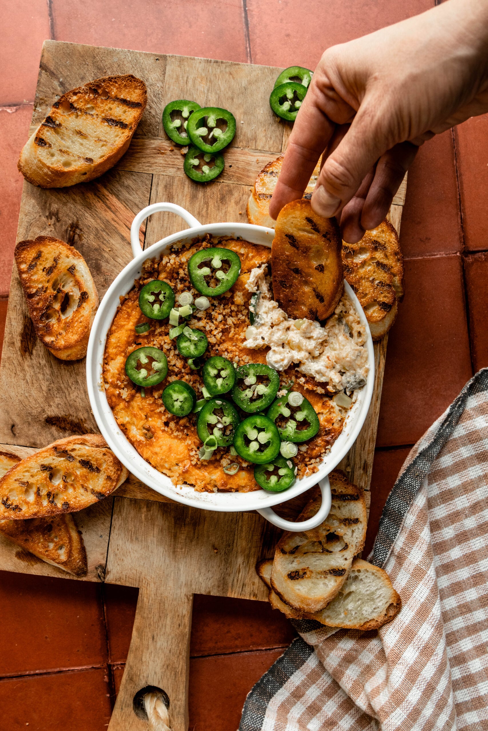 Alt text: Overhead shot of jalapeño chicken popper dip in a white baking dish, topped with sliced jalapeños and breadcrumbs, served with grilled crostini on a wooden board. A hand is dipping a slice of grilled bread into the creamy dip.