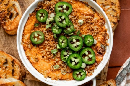 Alt text: Overhead shot of jalapeño chicken popper dip in a white baking dish, topped with sliced jalapeños, crispy panko breadcrumbs, and chopped green onions. Surrounded by grilled crostini on a wooden board over terracotta tile, with a checkered cloth in the corner.