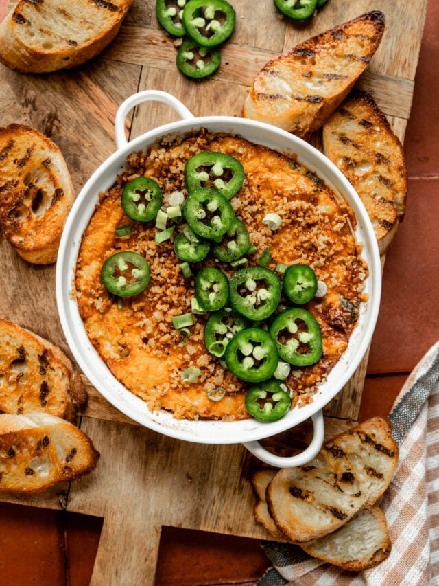 Alt text: Overhead shot of jalapeño chicken popper dip in a white baking dish, topped with sliced jalapeños, crispy panko breadcrumbs, and chopped green onions. Surrounded by grilled crostini on a wooden board over terracotta tile, with a checkered cloth in the corner.