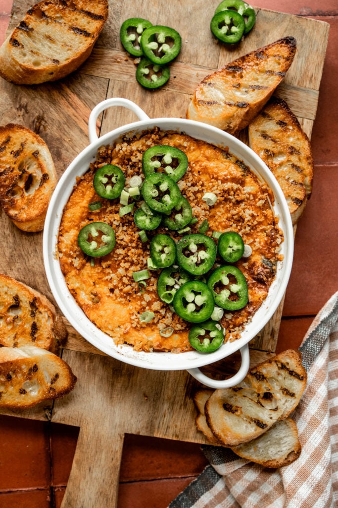 Alt text: Overhead shot of jalapeño chicken popper dip in a white baking dish, topped with sliced jalapeños, crispy panko breadcrumbs, and chopped green onions. Surrounded by grilled crostini on a wooden board over terracotta tile, with a checkered cloth in the corner.