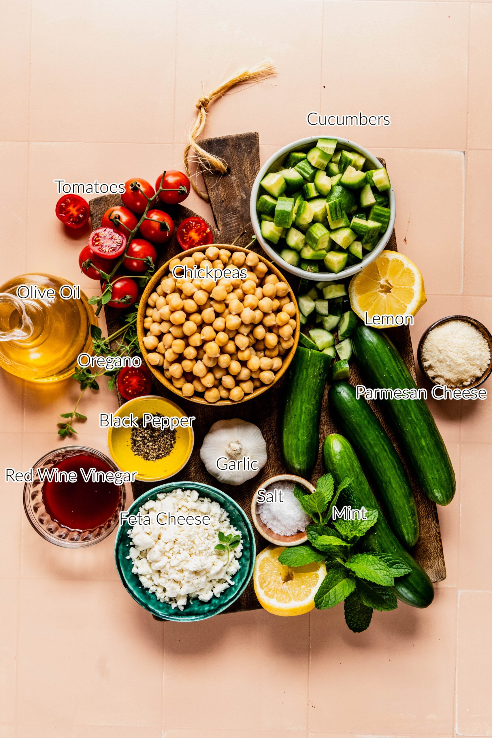 Flat lay of fresh ingredients for a cucumber, tomato, and feta salad arranged on a wooden board and pink tile surface. Labeled items include cherry tomatoes, chopped cucumbers, chickpeas, crumbled feta cheese, fresh mint, lemon, garlic, oregano, red wine vinegar, olive oil, Parmesan cheese, salt, and black pepper. The setup showcases vibrant, wholesome components used to make a Mediterranean-style salad.