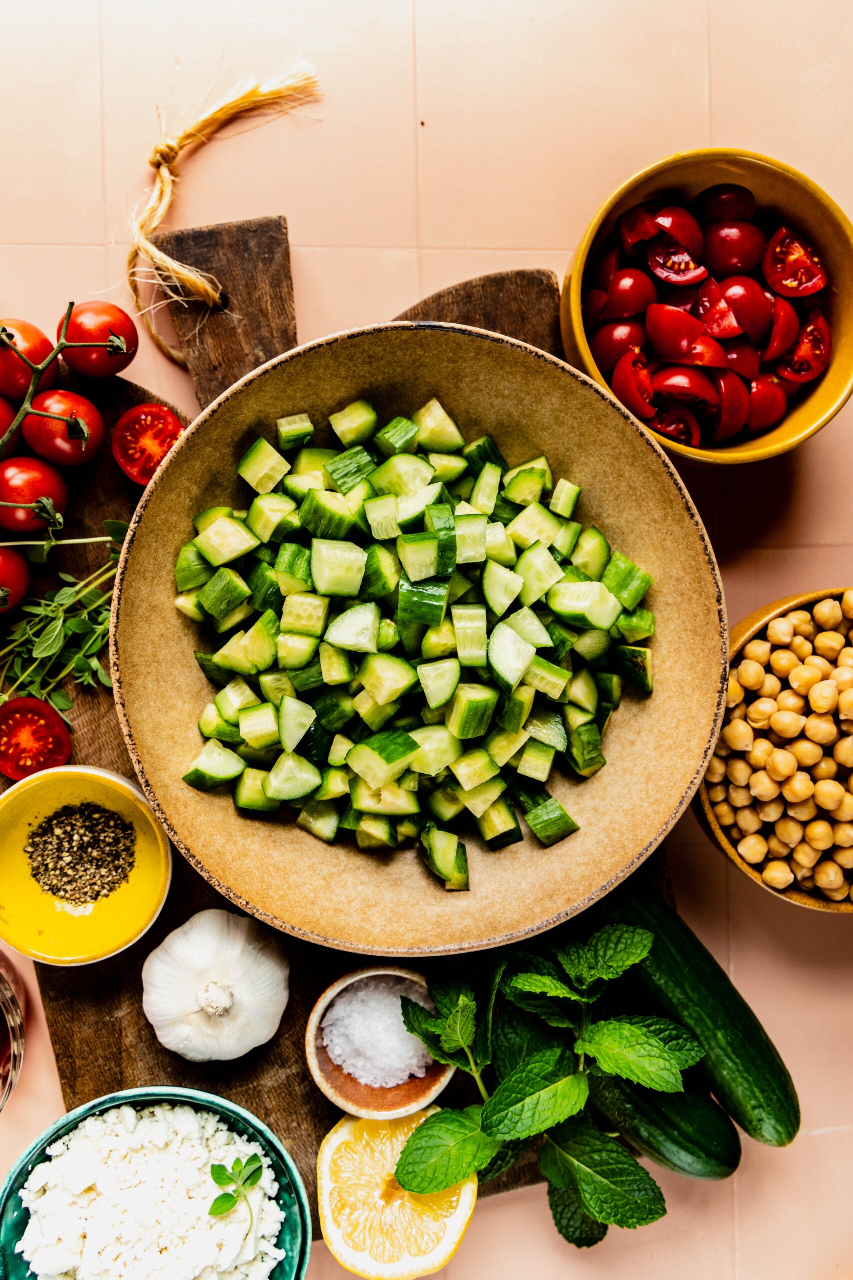 Overhead view of ingredients for a cucumber, tomato, and feta salad arranged on a wooden board. The centerpiece is a bowl filled with chopped cucumbers, surrounded by bowls of halved cherry tomatoes, chickpeas, crumbled feta cheese, cracked black pepper, garlic, fresh mint, salt, oregano, and a halved lemon. The setup highlights the fresh, vibrant components of a Mediterranean-style salad.