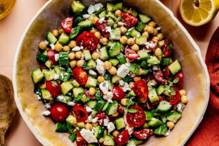**Alt Text:** Overhead view of a fresh cucumber, tomato, and feta salad served in a large beige bowl on a peach-colored tiled surface. The salad is surrounded by ingredients including whole and sliced cucumbers, cherry tomatoes on the vine, a halved lemon, a bottle of olive oil, and a glass of water. A red cloth napkin and wooden serving utensils complete the scene, evoking a bright and summery Mediterranean vibe.
