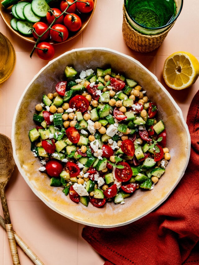 **Alt Text:** Overhead view of a fresh cucumber, tomato, and feta salad served in a large beige bowl on a peach-colored tiled surface. The salad is surrounded by ingredients including whole and sliced cucumbers, cherry tomatoes on the vine, a halved lemon, a bottle of olive oil, and a glass of water. A red cloth napkin and wooden serving utensils complete the scene, evoking a bright and summery Mediterranean vibe.