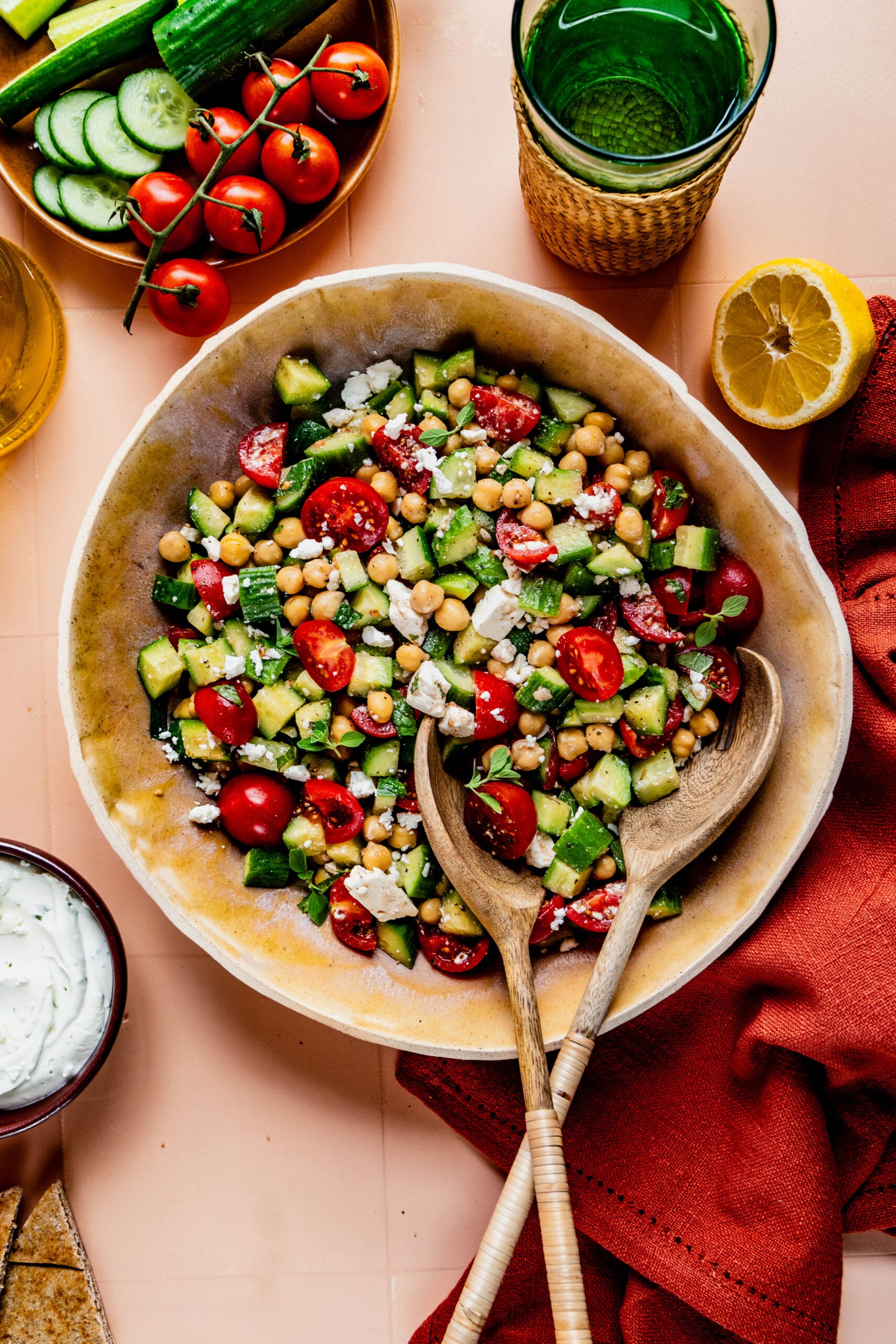 Overhead view of a vibrant cucumber, tomato, and feta salad served in a large ceramic bowl with wooden serving utensils. The salad features chopped cucumbers, halved cherry tomatoes, chickpeas, and crumbled feta, garnished with herbs. Surrounding the bowl are fresh ingredients including vine tomatoes, sliced cucumbers, a halved lemon, a glass of water, olive oil, and a small bowl of creamy dressing, set on a peach-colored surface with a red linen napkin.