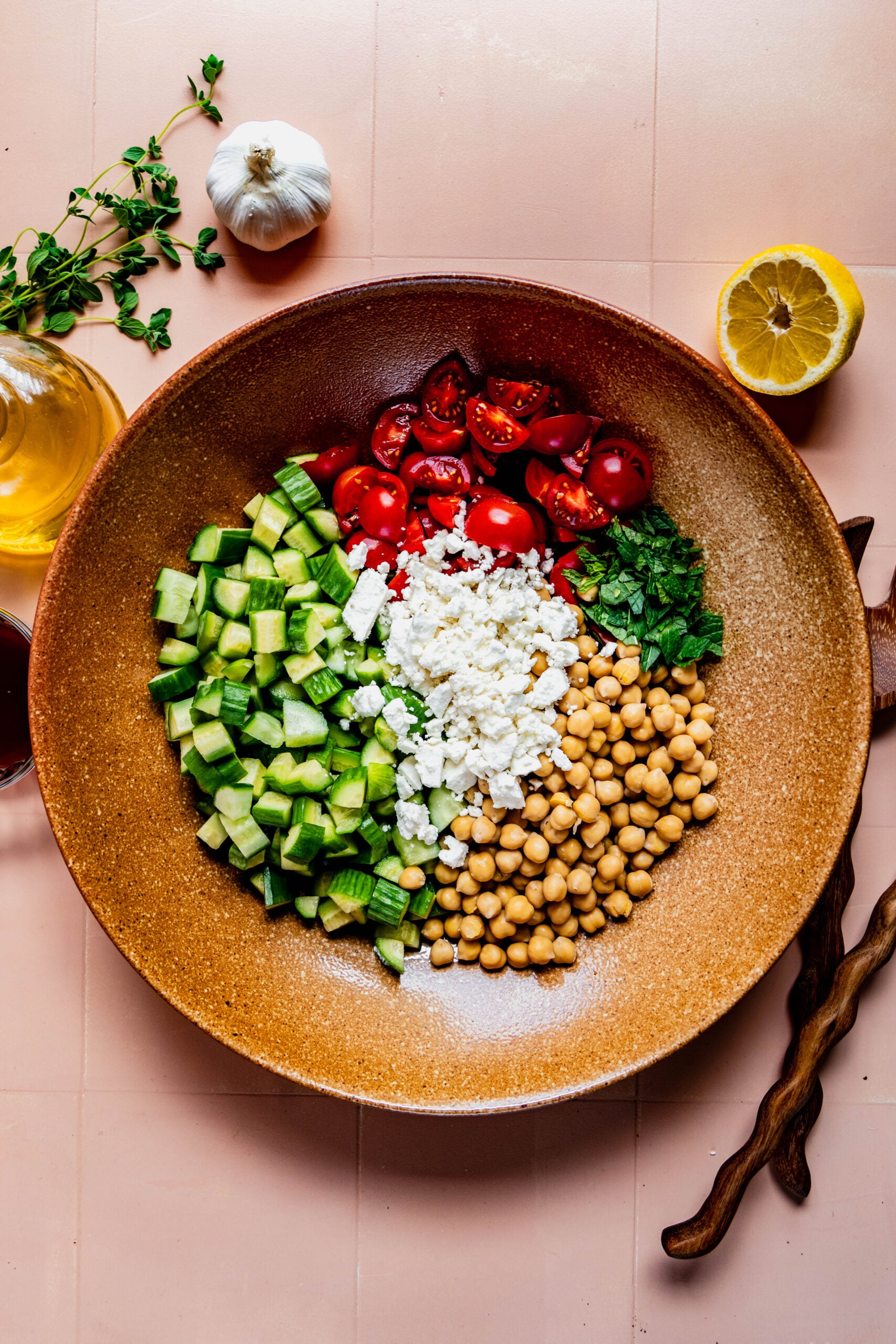 Overhead view of a vibrant cucumber, tomato, and feta salad in a large brown bowl. The salad features chopped cucumber, quartered cherry tomatoes, crumbled feta, chickpeas, and fresh mint arranged in neat sections. Surrounding the bowl are ingredients for the dressing: a halved lemon, garlic bulb, olive oil, red wine vinegar, and fresh oregano, all set on a pink tile background.