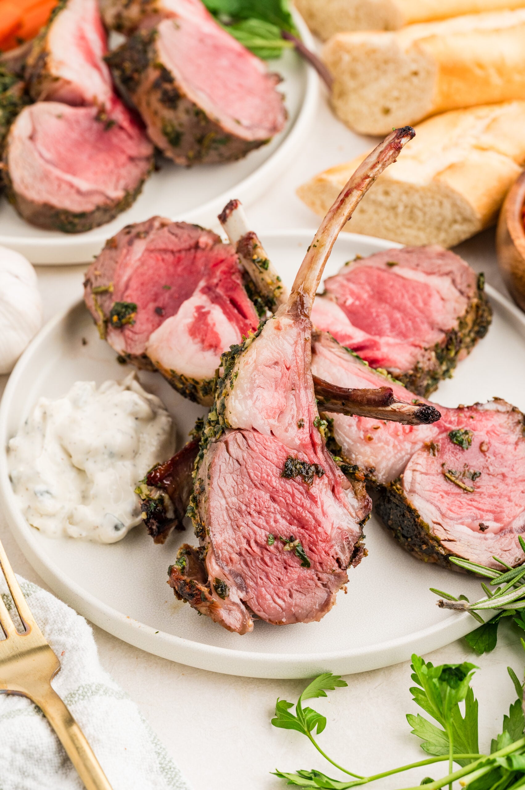 Sliced herb crusted rack of lamb recipe served on a white plate with a dollop of tzatziki sauce, garnished with fresh parsley and rosemary. Baguette slices and additional lamb pieces are visible in the background.