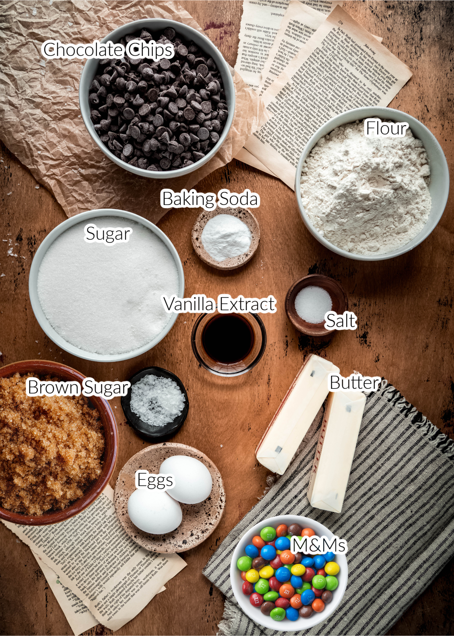 Flat lay of labeled ingredients for chewy M&M cookies, including bowls of flour, sugar, brown sugar, chocolate chips, M&Ms, baking soda, salt, vanilla extract, eggs, and sticks of butter arranged on a rustic wooden surface with recipe pages