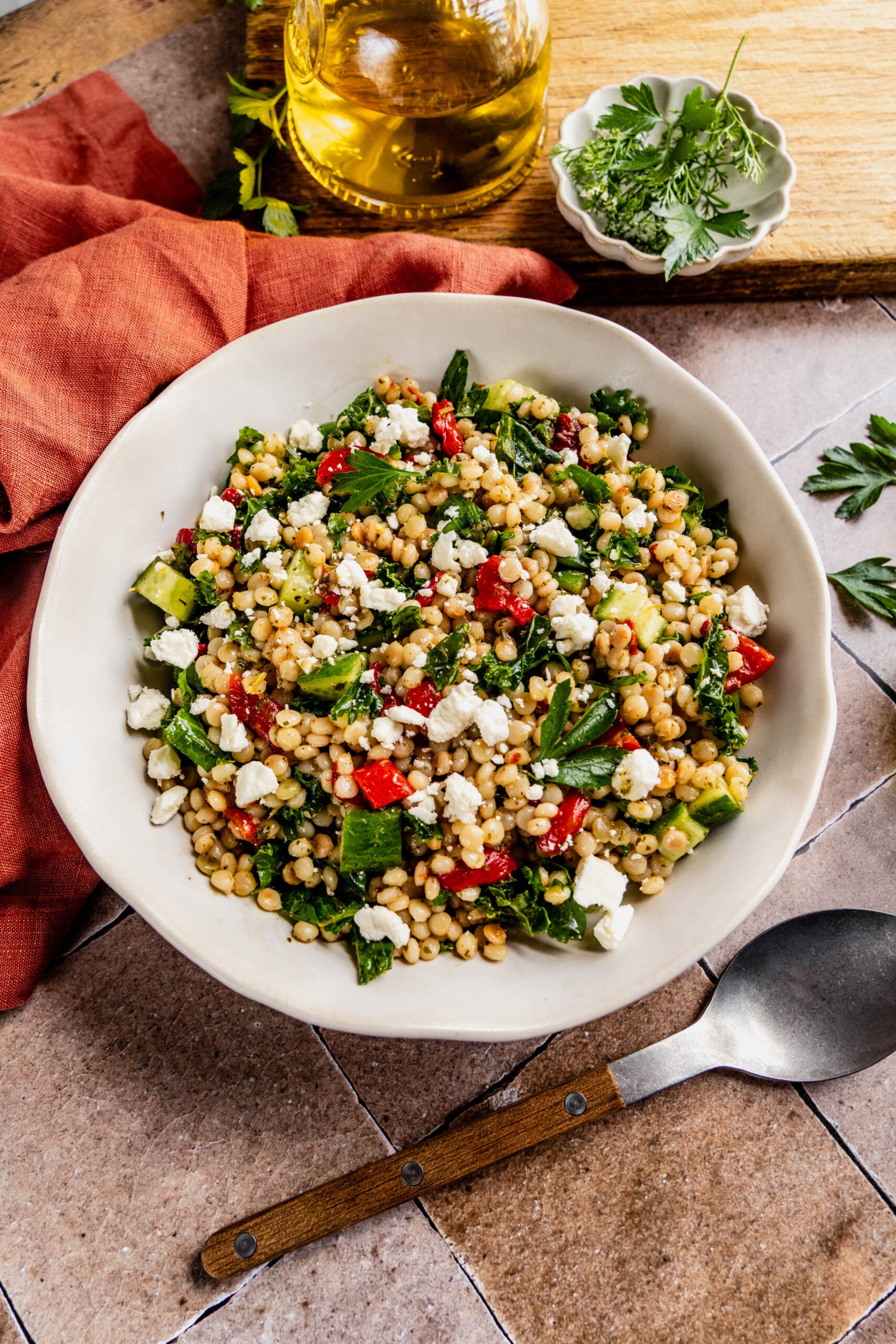 close up view of a vibrant couscous kale salad served in a white bowl with a wooden-handled fork, featuring fluffy couscous, chopped kale, diced cucumbers, roasted red peppers, and crumbled feta cheese. The scene includes a small bowl of fresh herbs, a jar of olive oil, all styled on a rustic tiled surface with a burnt orange linen napkin.