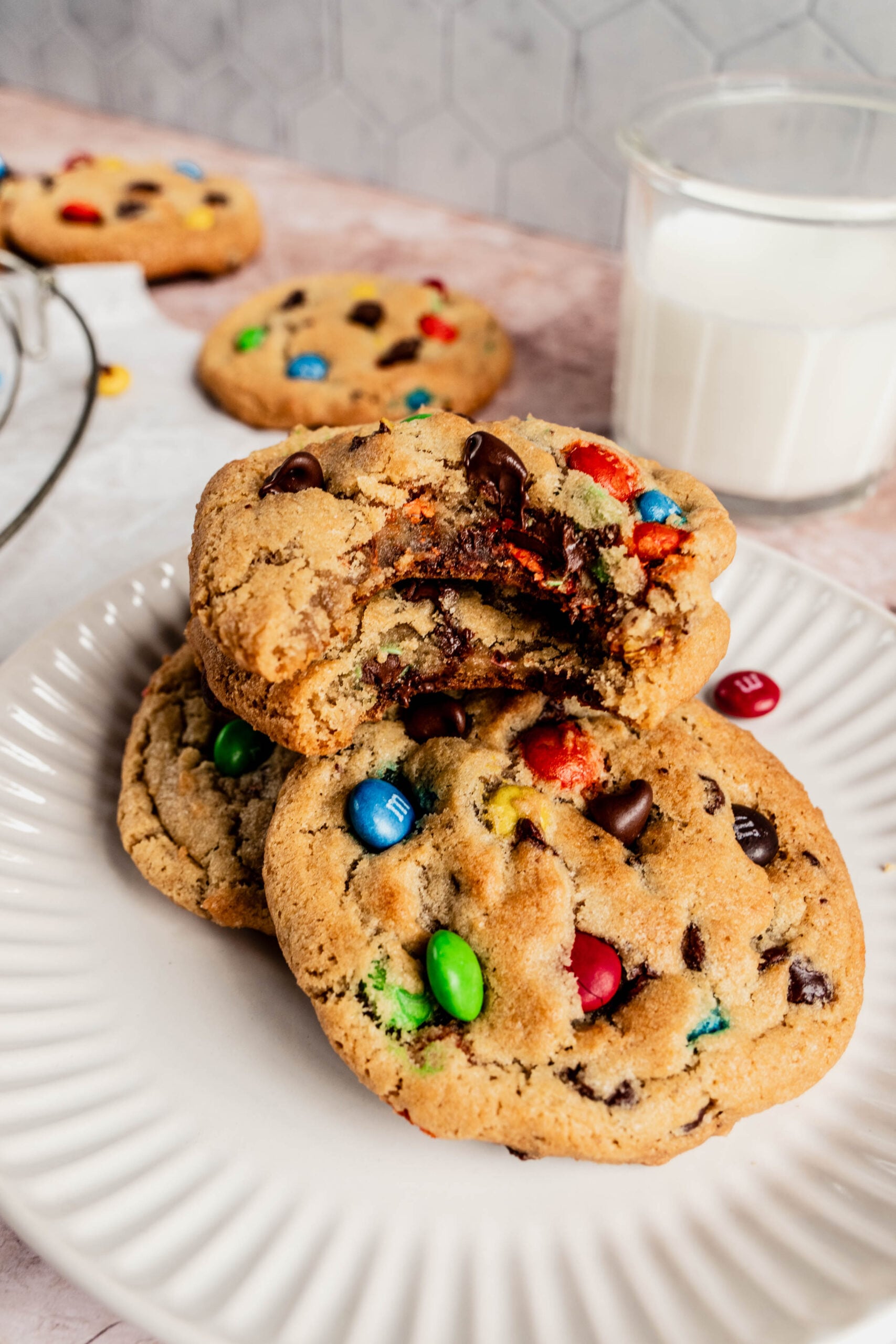 Close-up of chewy M&M cookies stacked on a white plate, with one cookie broken in half showing the gooey chocolate center and colorful candy pieces, served with a glass of milk in the background