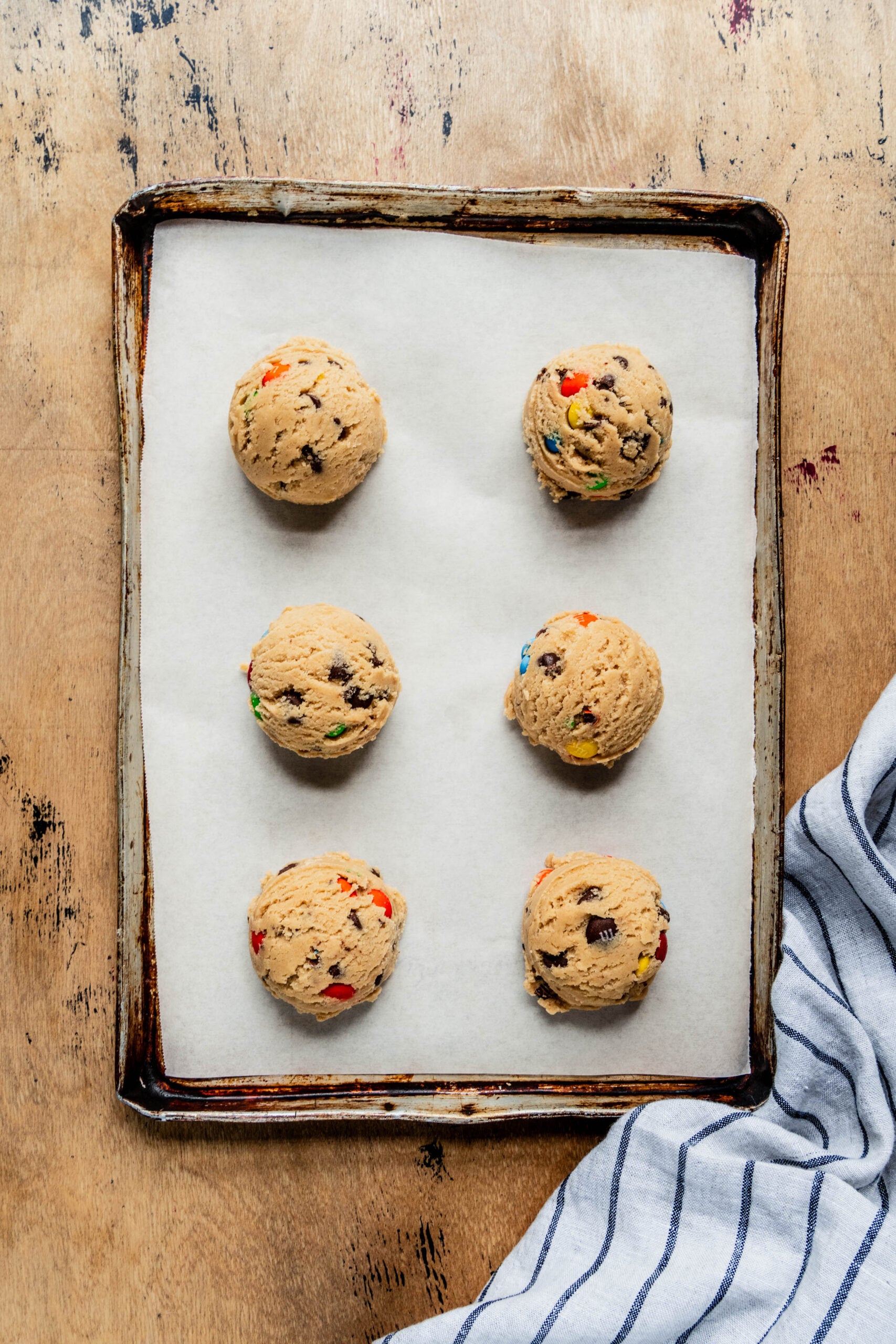 Six scoops of chewy M&M cookie dough arranged on a parchment-lined baking sheet, ready to be baked, with colorful candy pieces and chocolate chips visible in the dough.