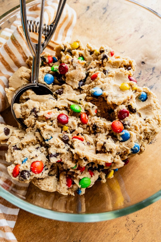 A glass bowl filled with chewy M\&M cookie dough mixed with colorful candy-coated chocolates and chocolate chips, with a metal cookie scoop resting inside, placed on a wooden surface with a striped kitchen towel.
