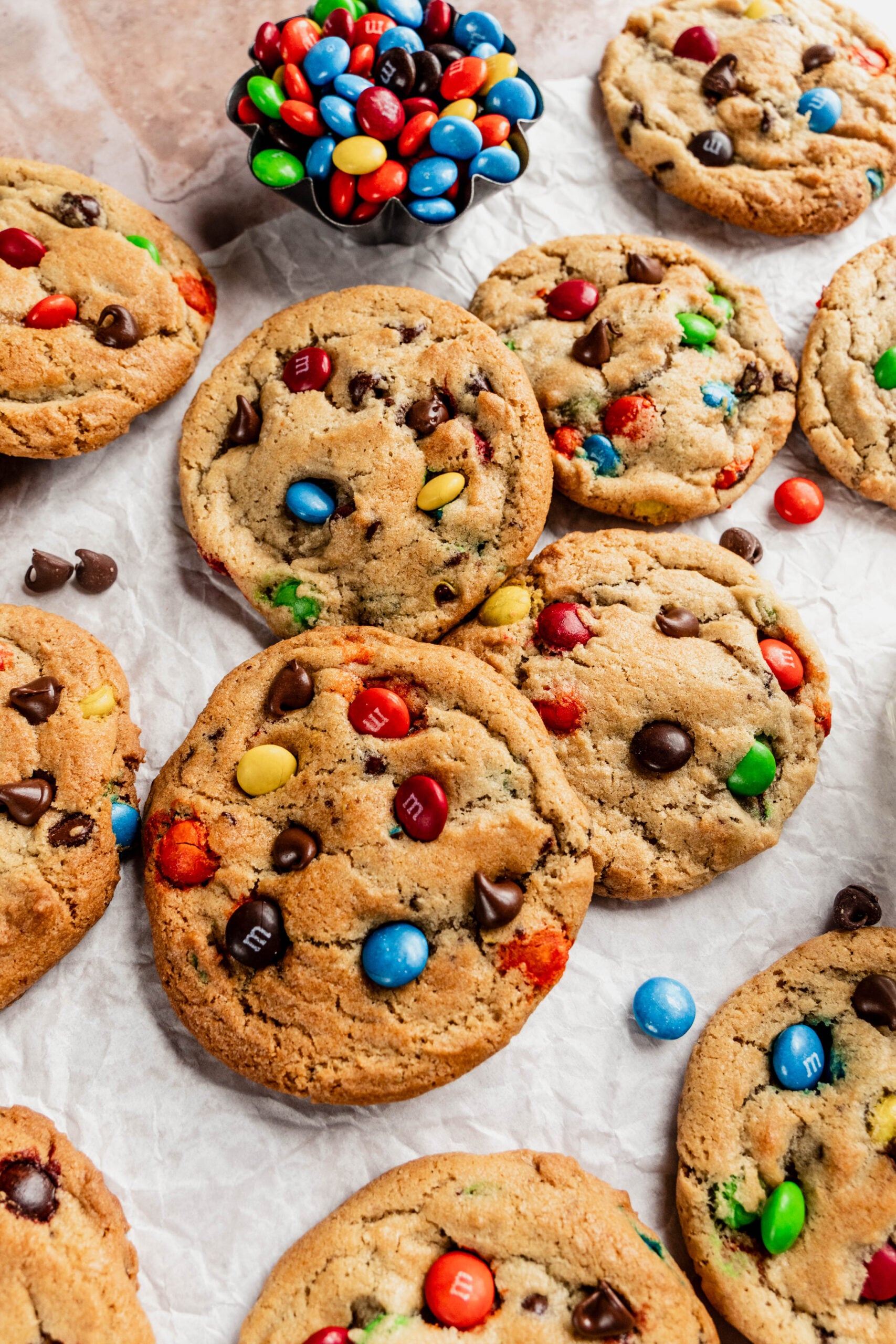 Freshly baked chewy M&M cookies loaded with colorful candy-coated chocolates and chocolate chips, arranged on parchment paper with a small bowl of M&Ms in the background.