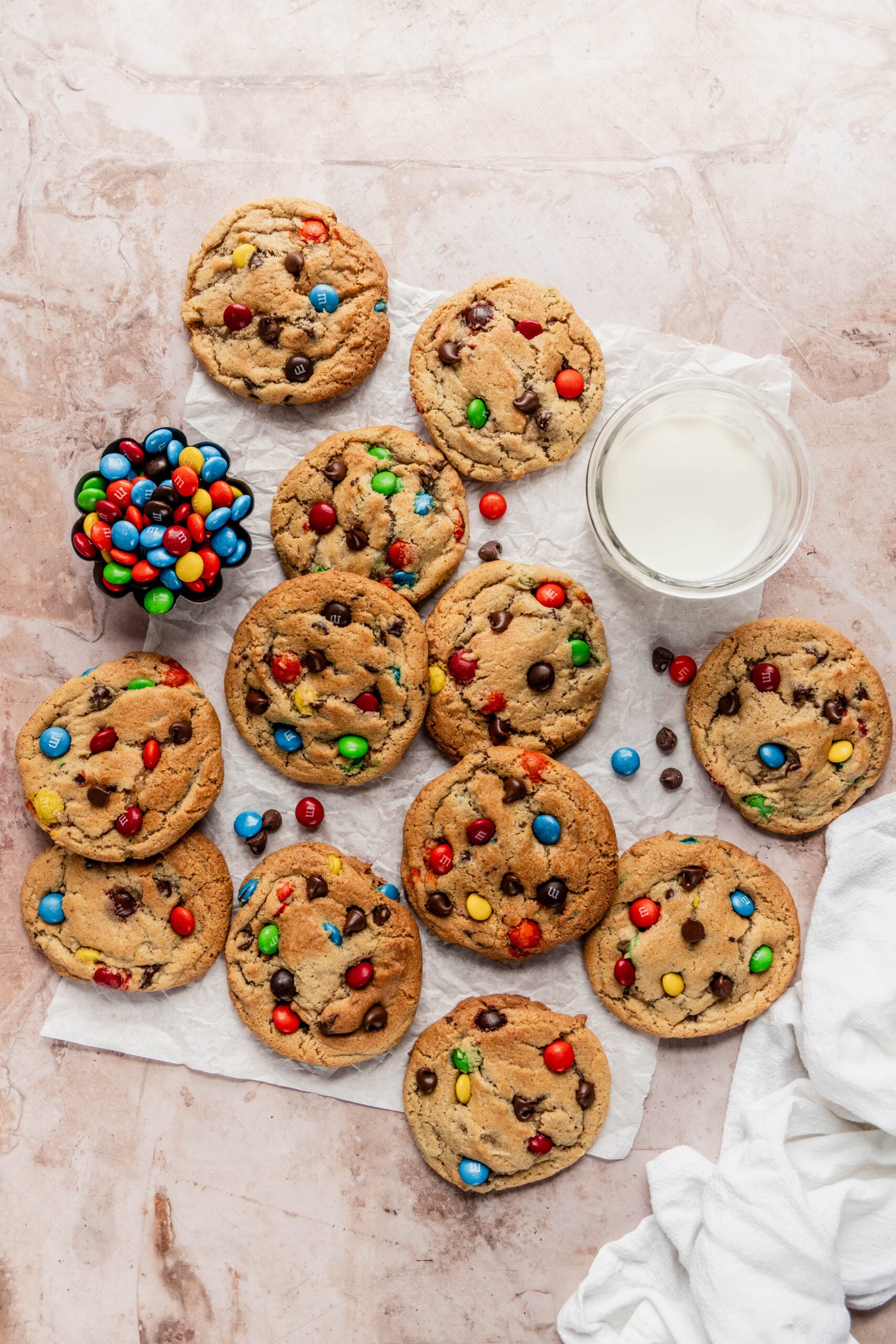Overhead view of a batch of chewy M&M cookies with colorful candy pieces and chocolate chips, arranged on parchment paper next to a glass of milk and a small bowl of M&Ms