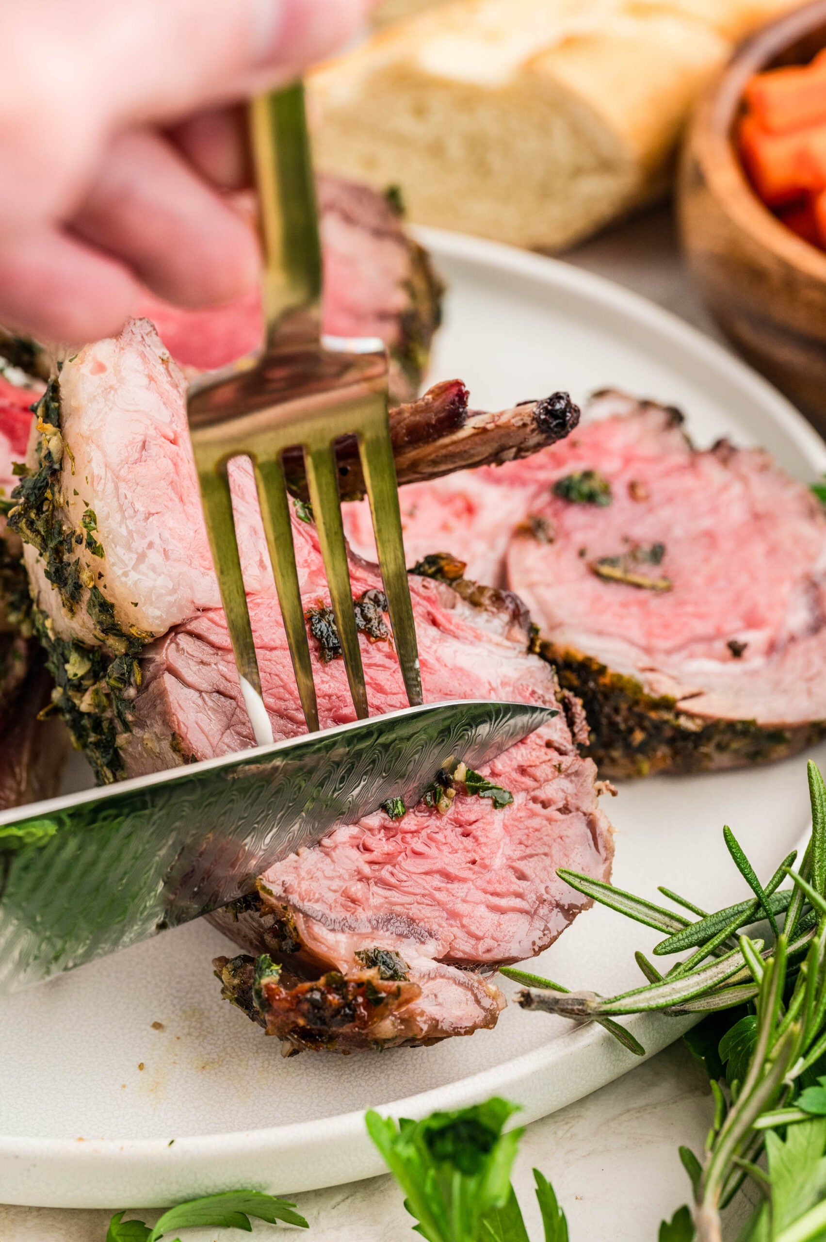 Close-up of herb crusted rack of lamb recipe being sliced with a knife and held steady with a fork, revealing tender, juicy medium-rare meat. Fresh rosemary and parsley are scattered on the plate with bread and vegetables in the background.