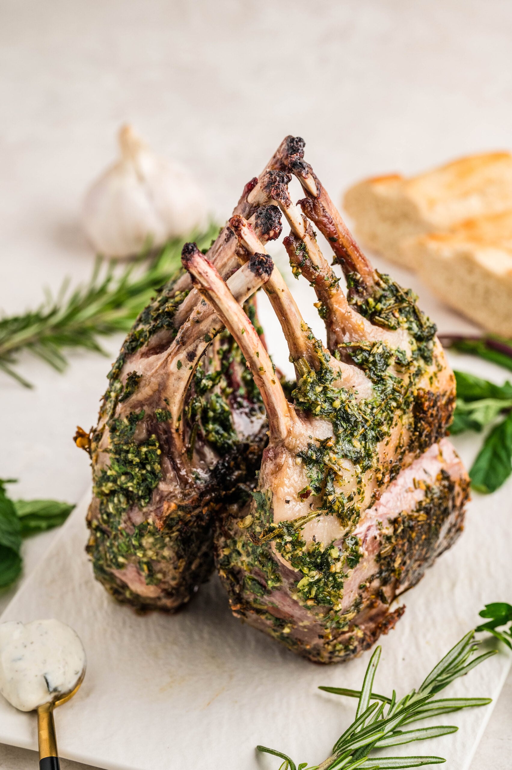 Close-up of herb crusted rack of lamb recipe, standing upright with bones interlaced, coated in a vibrant green herb and garlic mixture. Fresh rosemary, mint, and a spoonful of tzatziki sauce are arranged around it on a white surface.