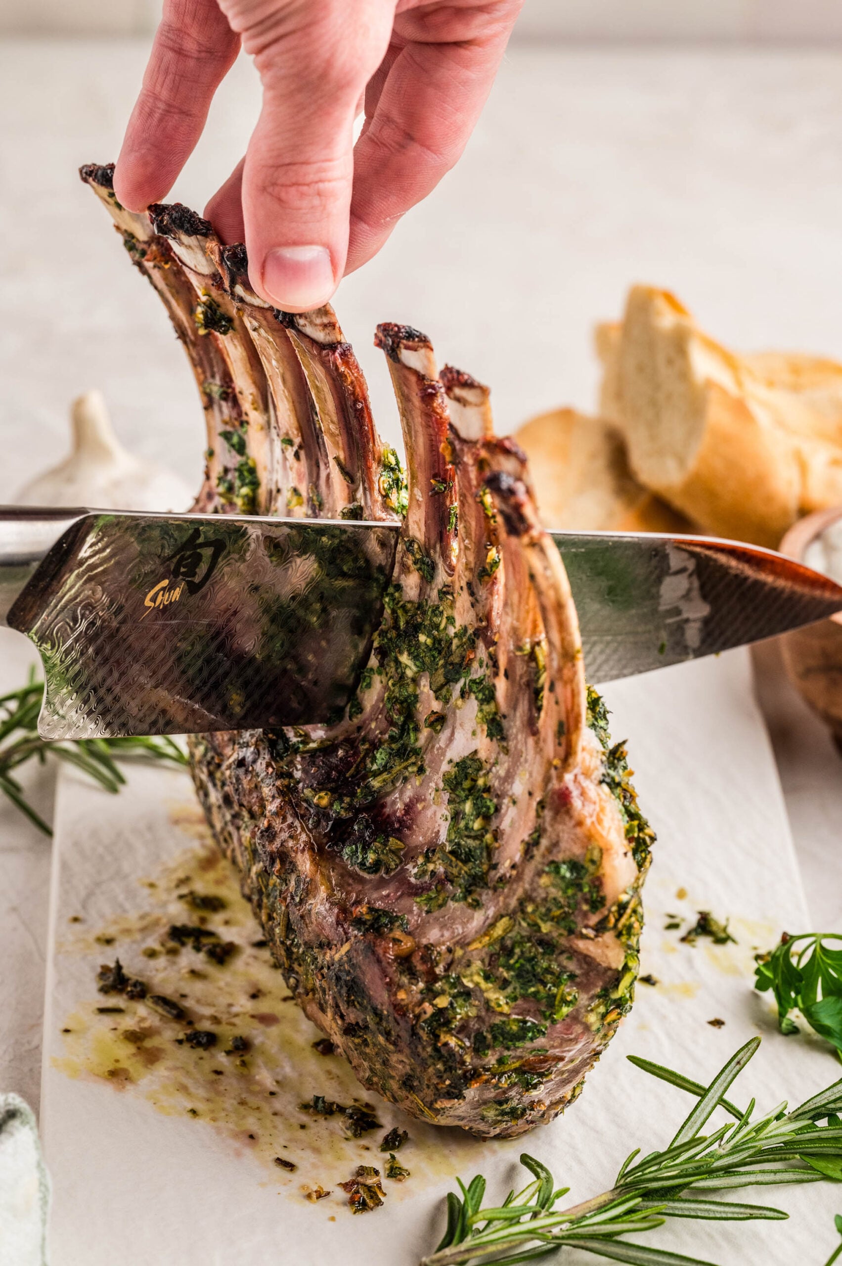 Close-up of herb crusted rack of lamb recipe being sliced with a sharp knife while held upright by the bones. The lamb is coated in a fresh herb and garlic crust, with rosemary sprigs and bread in the background.