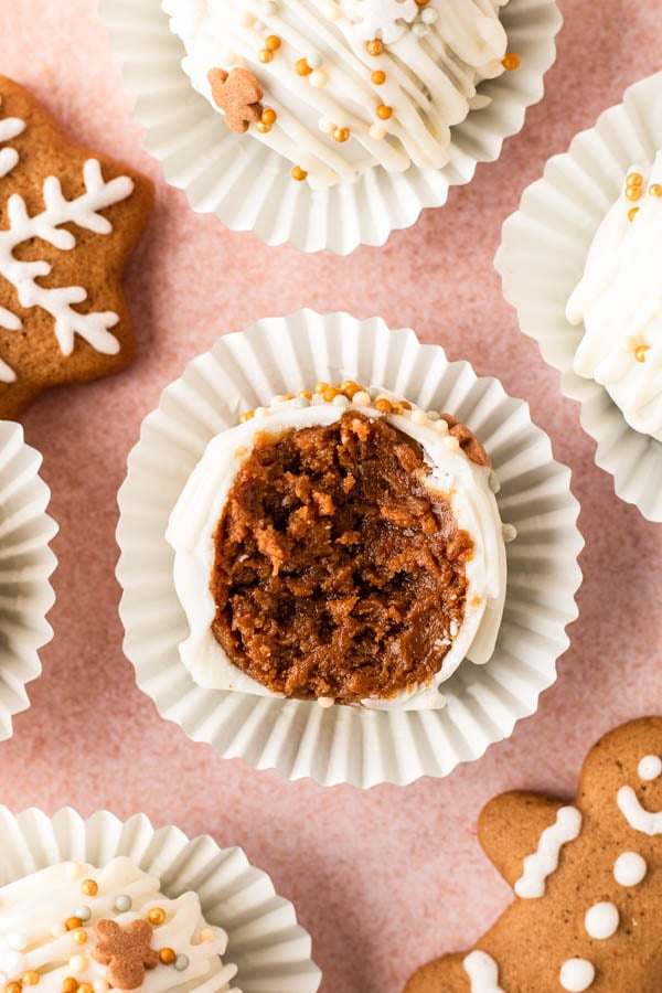 Close-up of gingerbread truffles coated in white chocolate, drizzled with more chocolate, and topped with festive gold and white sprinkles. One truffle is cut in half to reveal a rich, spiced gingerbread filling inside, all arranged in white paper liners on a serving plate with gingersnap cookies nearby.
