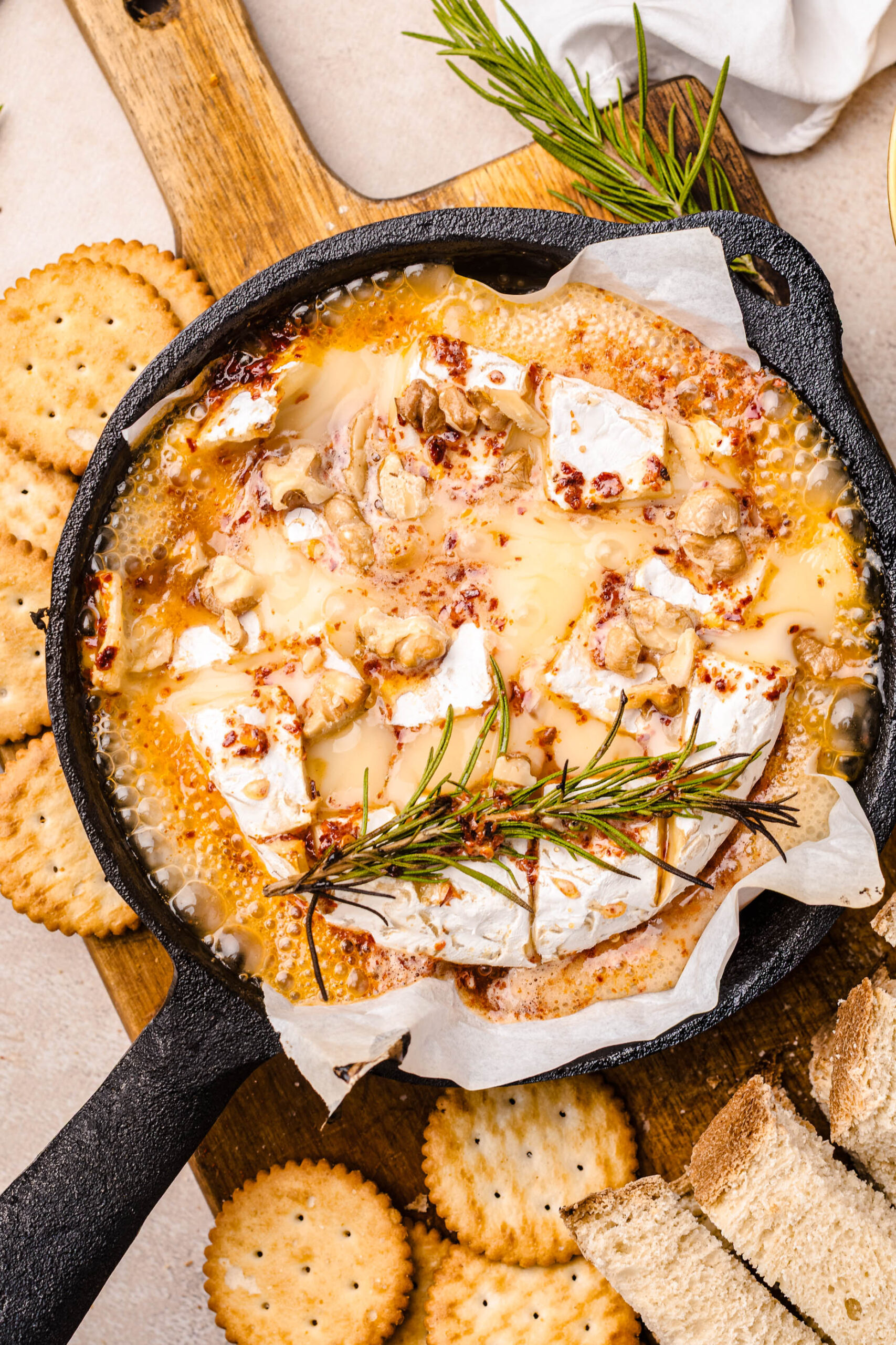 Overhead view of a baked Camembert recipe in a small cast-iron skillet, topped with bubbling hot honey, toasted walnuts, red pepper flakes, and a sprig of fresh rosemary. The melted cheese is surrounded by round buttery crackers and rustic bread slices for dipping, all arranged on a wooden serving board for a cozy, rustic appetizer presentation.