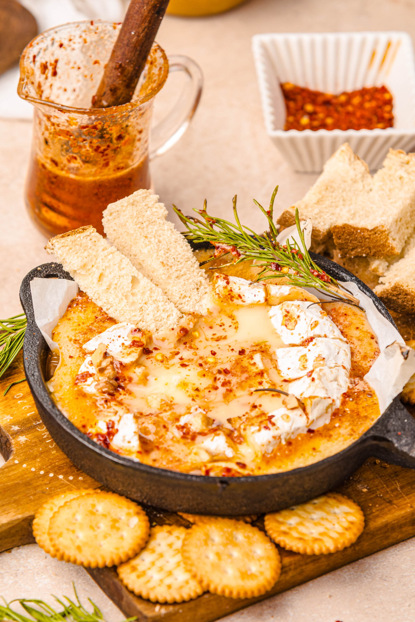 Warm baked Camembert served in a cast-iron skillet, topped with bubbling hot honey, red pepper flakes, toasted walnuts, and a sprig of rosemary. The melted cheese is surrounded by toasted bread slices and round crackers, with a glass pitcher of spicy honey in the background, creating a cozy and inviting appetizer scene.