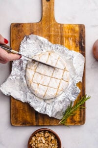 Overhead view of a wheel of Camembert cheese on a piece of foil atop a wooden cutting board, being scored in a crisscross pattern with a knife. A sprig of fresh rosemary and a small bowl of chopped walnuts sit nearby, showing preparation steps for a baked Camembert recipe.