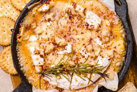 Overhead view of a baked Camembert recipe in a small cast-iron skillet, topped with bubbling hot honey, toasted walnuts, red pepper flakes, and a sprig of fresh rosemary. The melted cheese is surrounded by round buttery crackers and rustic bread slices for dipping, all arranged on a wooden serving board for a cozy, rustic appetizer presentation.