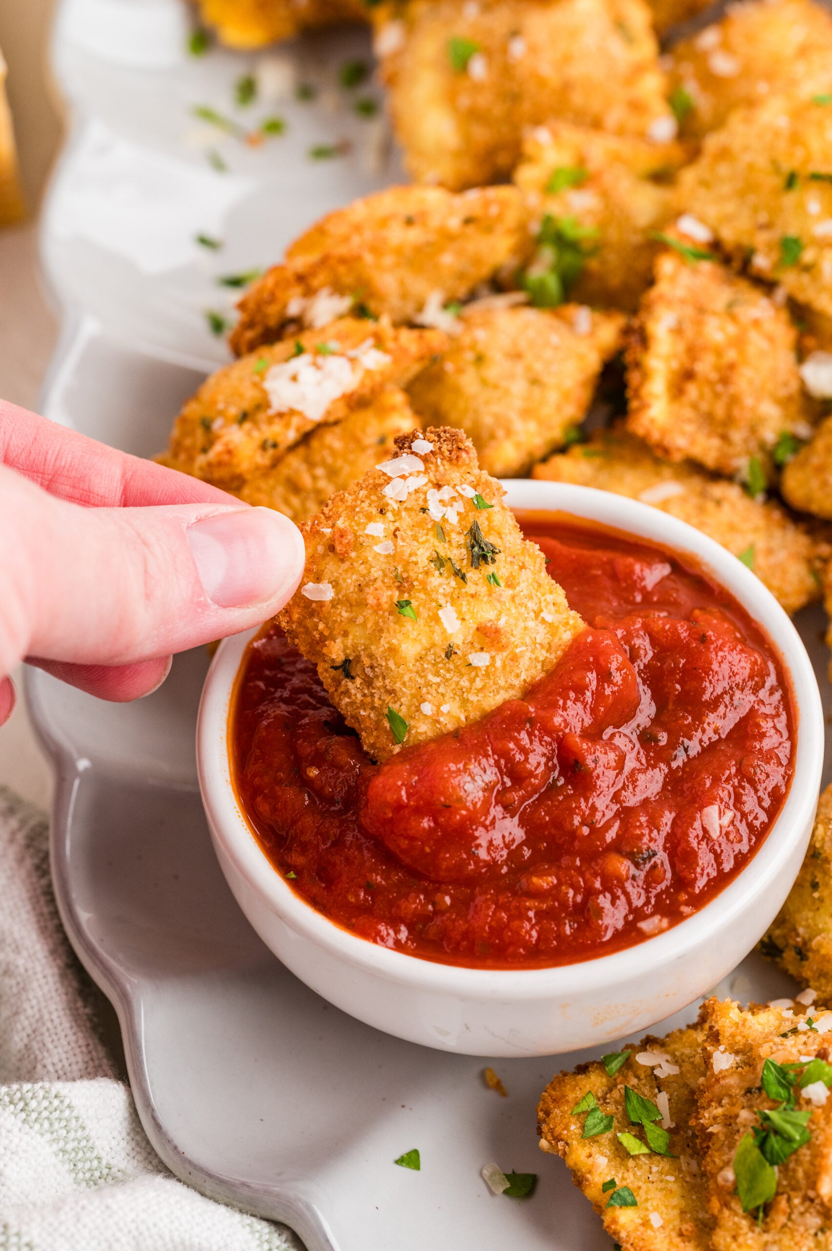 Close-up of a crispy air fryer toasted ravioli being dipped into a small bowl of warm marinara sauce, with more golden breaded ravioli sprinkled with parmesan and parsley in the background.