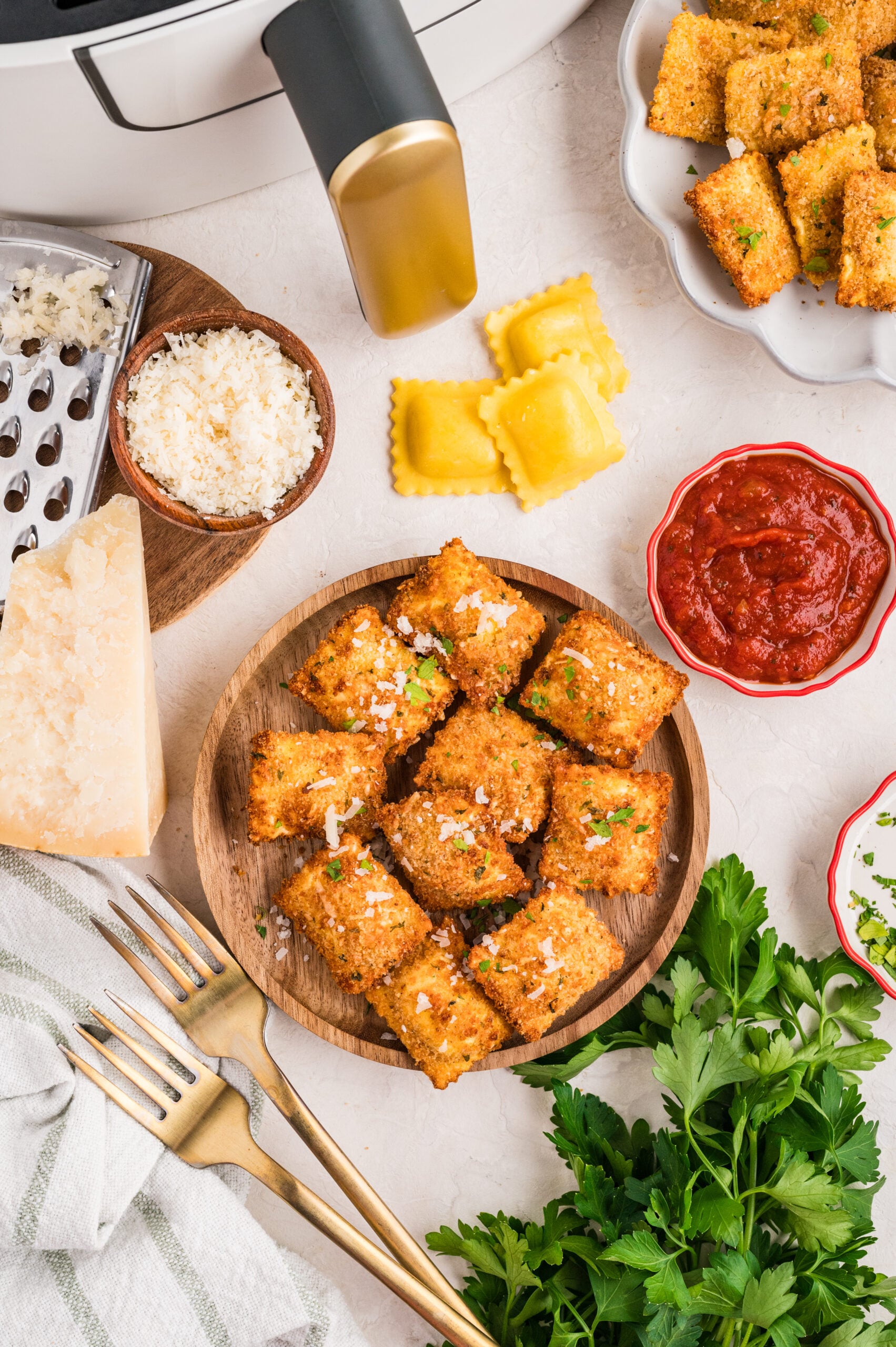 Overhead view of air fryer toasted ravioli served on a wooden plate, garnished with parmesan and parsley, surrounded by fresh ravioli, marinara sauce, grated cheese, herbs, and an air fryer in the background.