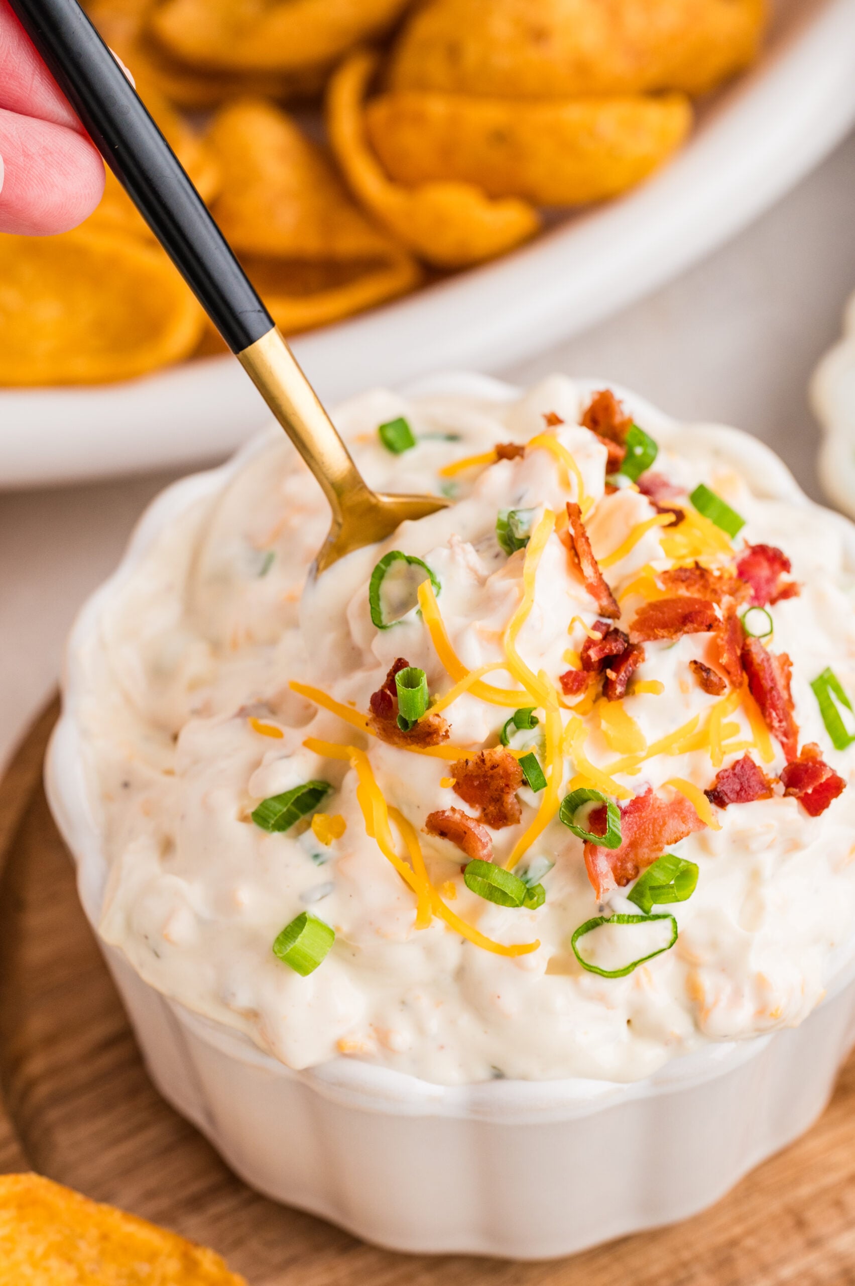 Close-up of creamy bacon ranch cheddar dip in a white ramekin, topped with shredded cheddar cheese, crispy bacon pieces, and chopped green onions, with a gold and black spoon scooping up a bite and corn chips in the background.