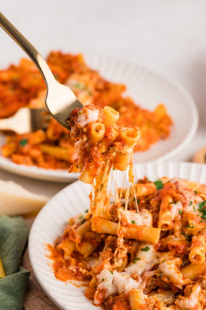 A fork lifting a cheesy bite of no boil baked ziti from a white plate, showing melted mozzarella stretching between tender pasta and rich tomato sauce, with another serving of ziti in the background.