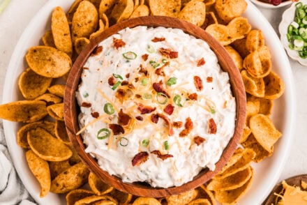 A wooden bowl filled with creamy bacon ranch cheddar dip topped with crispy bacon crumbles, shredded cheddar cheese, and chopped green onions, surrounded by scoops-style corn chips on a white platter. Small bowls of extra bacon, cheese, and green onions are arranged nearby for garnish.