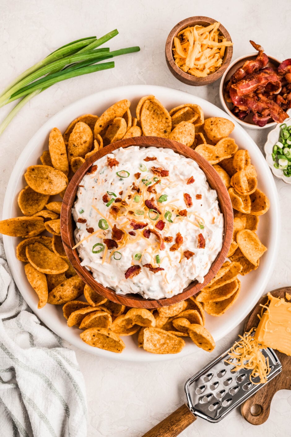 A wooden bowl filled with creamy bacon ranch cheddar dip topped with crispy bacon crumbles, shredded cheddar cheese, and chopped green onions, surrounded by scoops-style corn chips on a white platter. Small bowls of extra bacon, cheese, and green onions are arranged nearby for garnish.