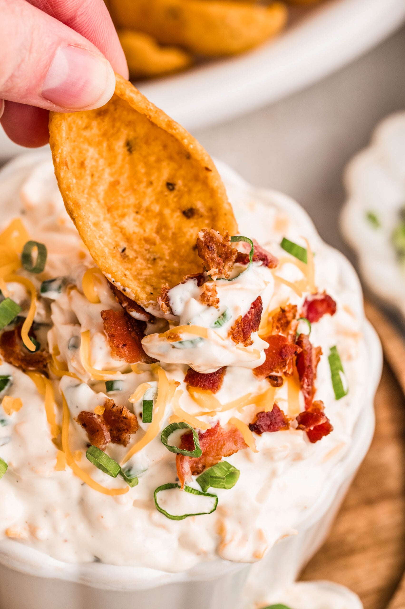 Close-up of a hand holding a corn chip dipping creamy bacon ranch cheddar dip in a white ramekin, topped with shredded cheddar cheese, crispy bacon pieces, and chopped green onions, with a gold and black spoon scooping up a bite and corn chips in the background.