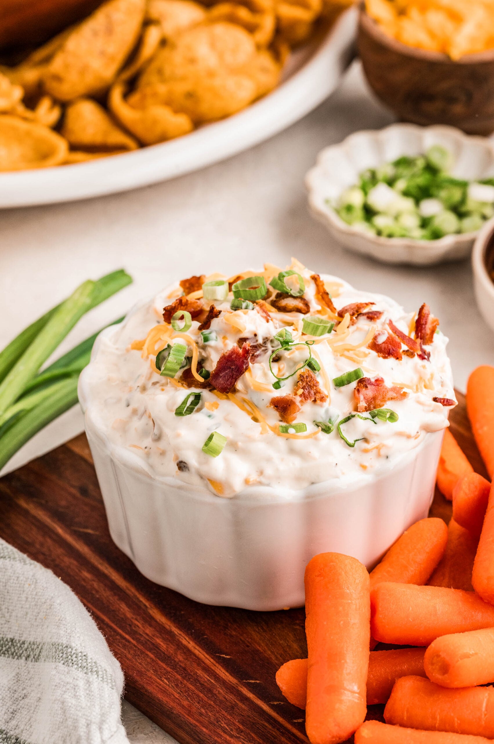 Close-up of creamy bacon ranch cheddar dip in a white ramekin, topped with shredded cheddar cheese, crispy bacon pieces, and chopped green onions, with a gold and black spoon scooping up a bite and corn chips in the background.