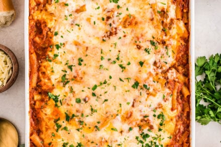 A large white casserole dish filled with golden, bubbly no boil baked ziti topped with melted mozzarella and sprinkled with fresh parsley. The dish is surrounded by ingredients including uncooked ziti, grated parmesan, fresh herbs, and sliced baguette on a light countertop.